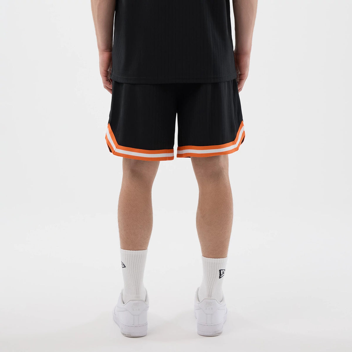 New Era New York Mets MLB Arch Black Oversized Shorts, back view on a model highlighting the relaxed fit with distinctive orange double stripe at the hemline; paired with white socks featuring an embroidered New Era logo above clean white trainers.