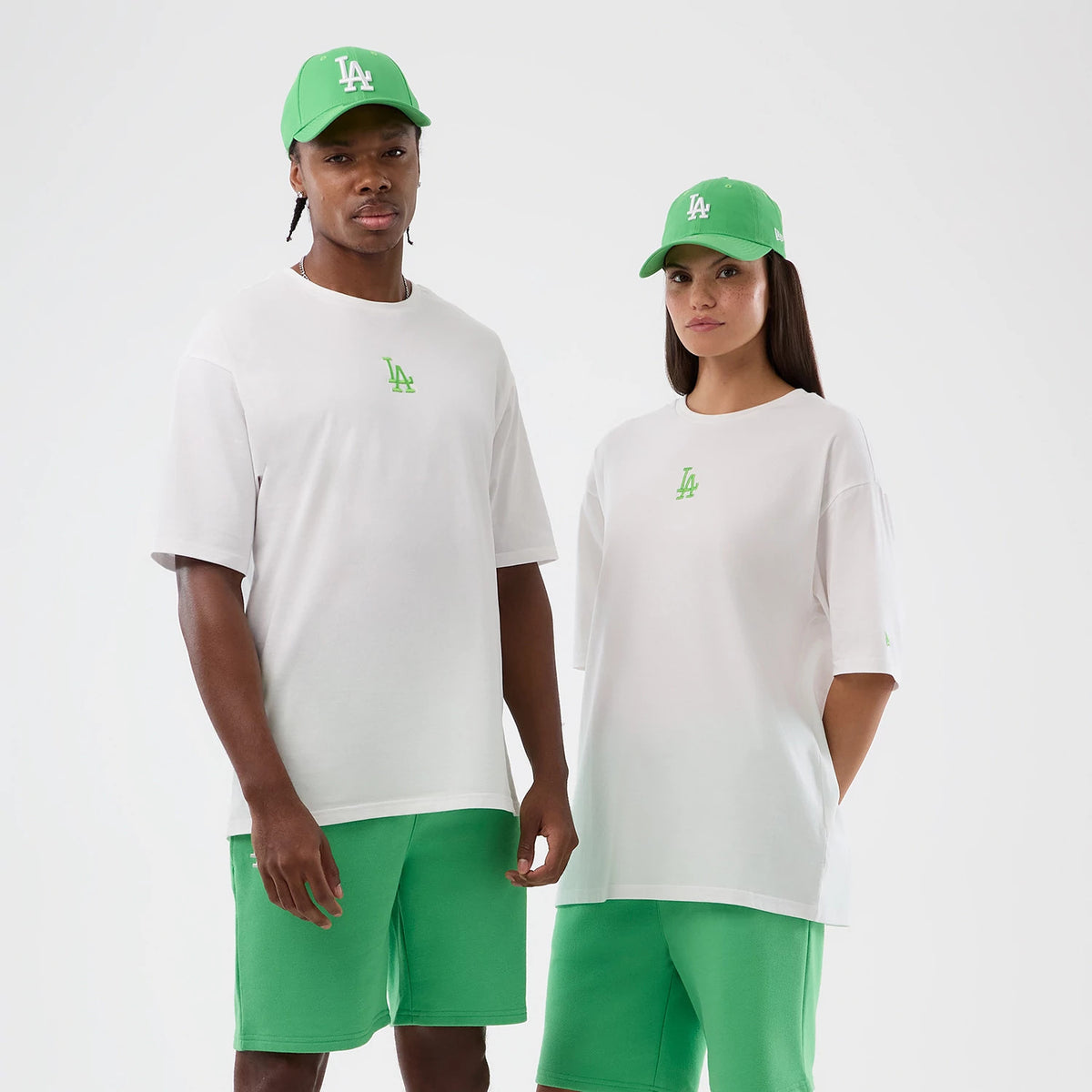 New Era LA Dodgers MLB League Essential White Oversized T-Shirt, front view on models wearing green caps and shorts, showing a large LA logo embroidered in green on the chest and the relaxed fit with short sleeves.