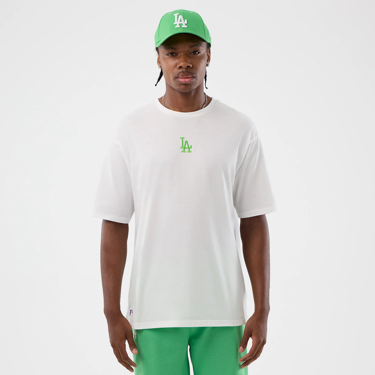New Era LA Dodgers MLB League Essential White Oversized T-Shirt, front view on a model styled with a matching green cap and trousers, featuring a green embroidered LA Dodgers logo centred on the chest against a plain white backdrop.