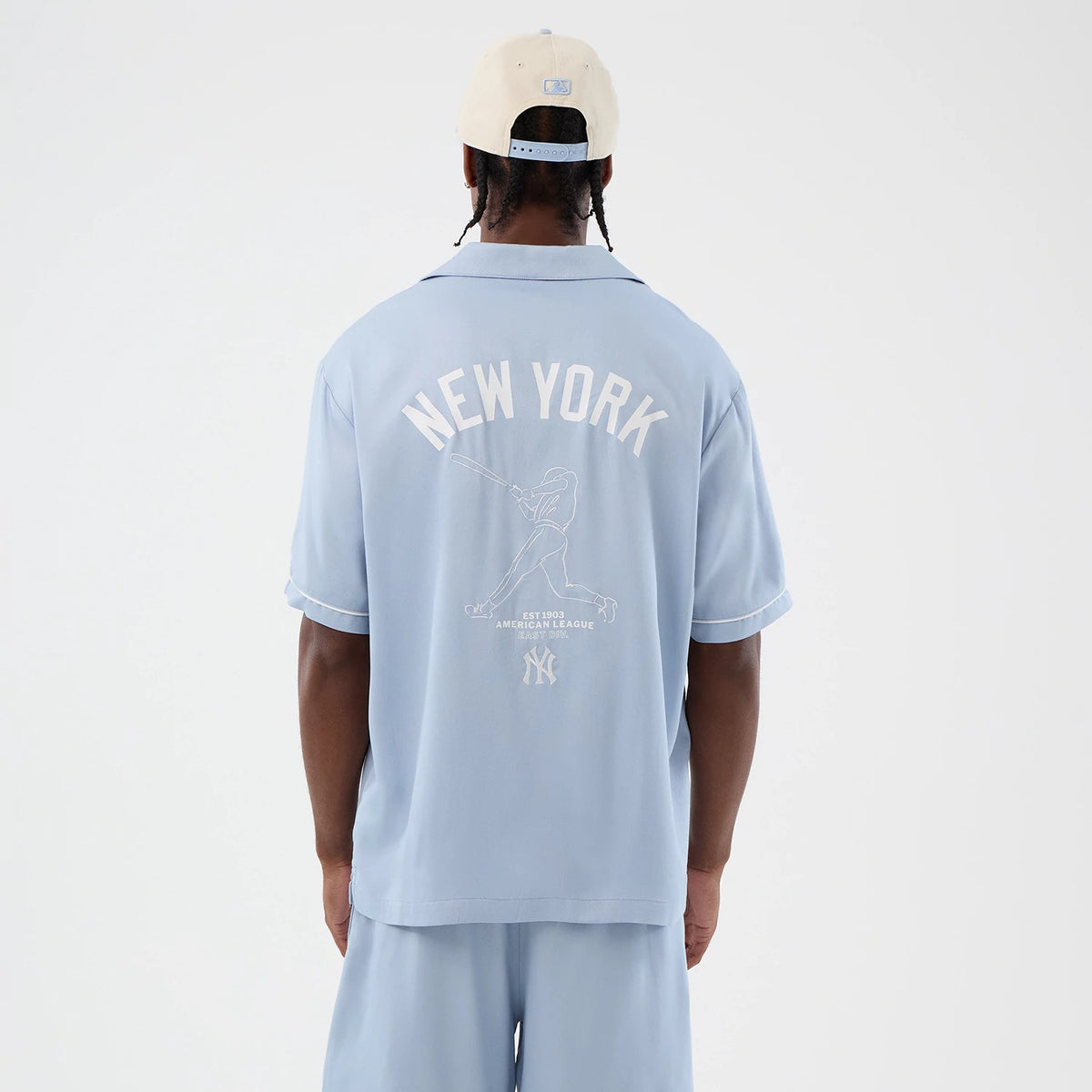 New Era New York Yankees MLB Graphic Revere Pastel Blue Short Sleeve Shirt, back view on a model wearing a cream cap, showing the large white NEW YORK lettering and a baseball player graphic, with a Yankees logo and piping on the sleeves.