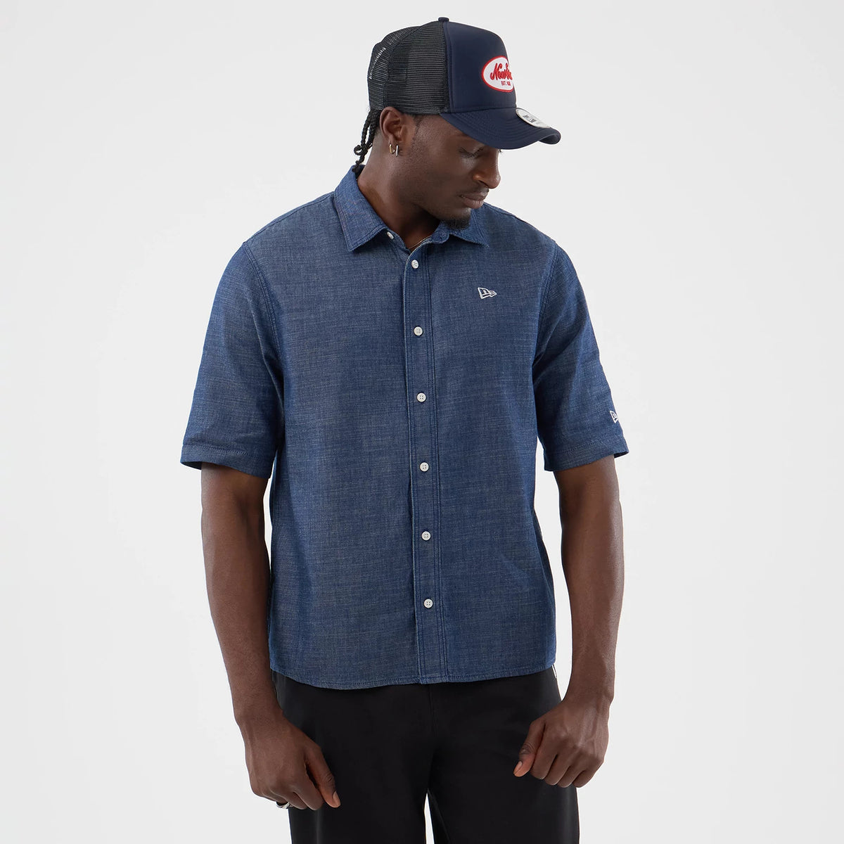 New Era Denim Blue Short Sleeve Shirt, front view on a model featuring a minimalist chambray fabric with a structured collar, the New Era flag logo embroidered on the left chest, and button closures running down the front.