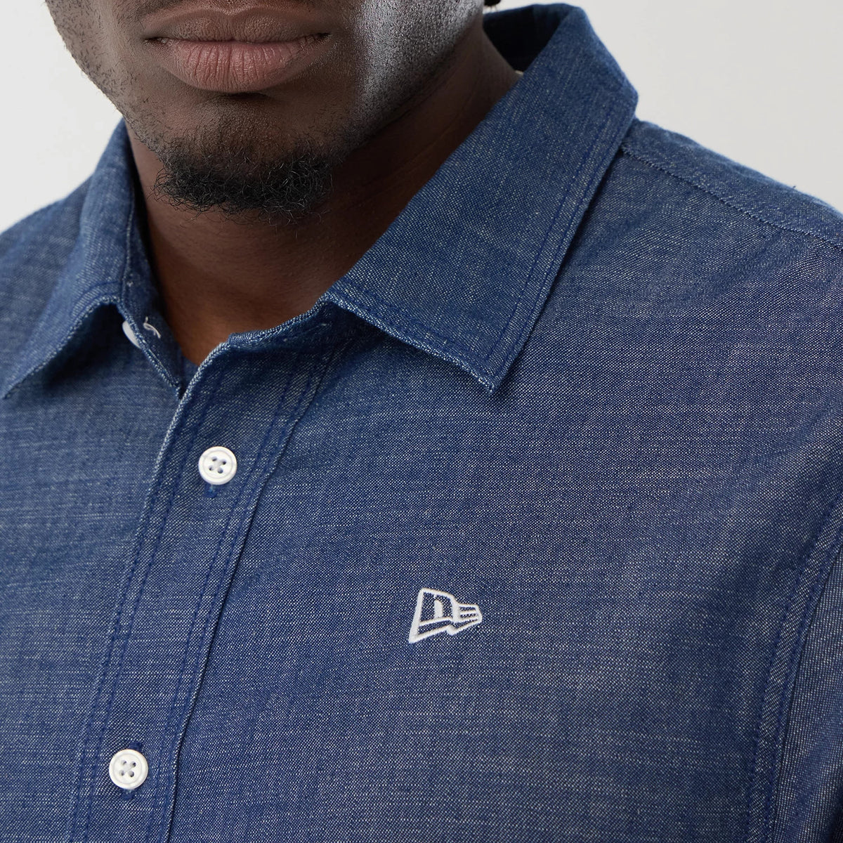 New Era Denim Blue Short Sleeve Shirt, close-up view showing white buttons on the front and a white embroidered New Era flag logo on the chest, with a classic collar and a fine denim texture.