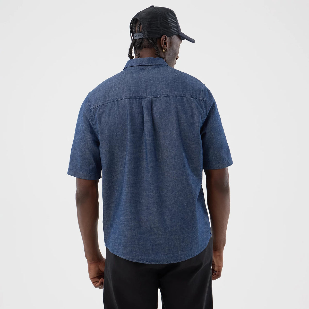New Era Denim Blue Short Sleeve Shirt, back view on a model with a black mesh cap, showing the shirt's straight hemline, relaxed fit, and a single pleat beneath the yoke for added style.