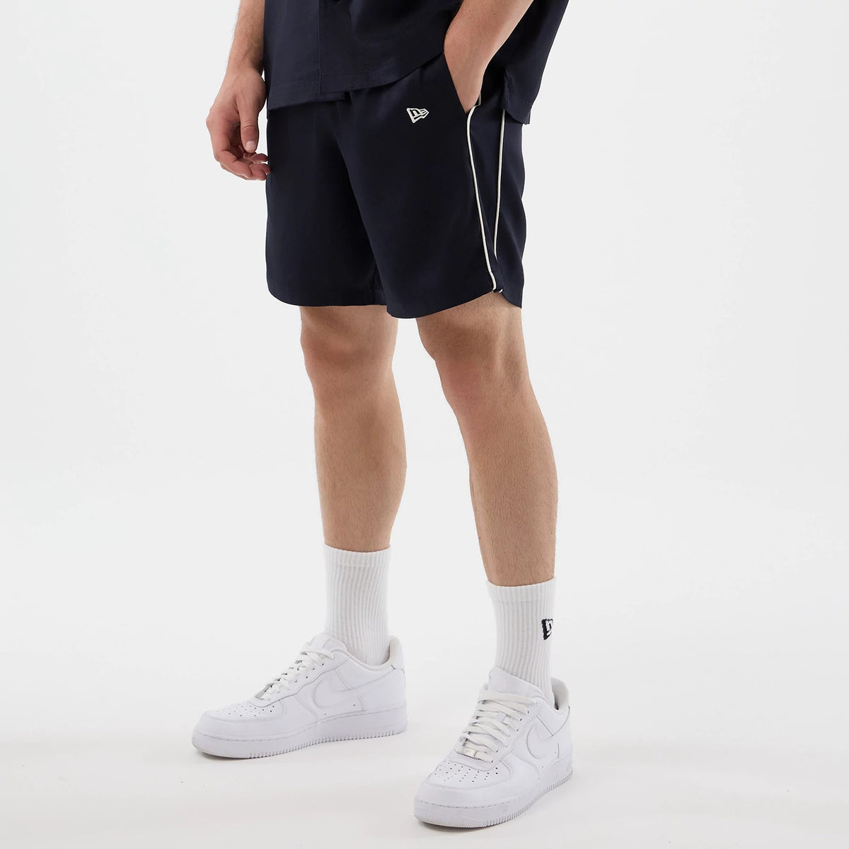New Era Piping Navy Shorts, on-model view showing the navy shorts with white piping along the side seams, an embroidered New Era logo on the left thigh, with the model wearing white trainers and socks, highlighting the casual styling.