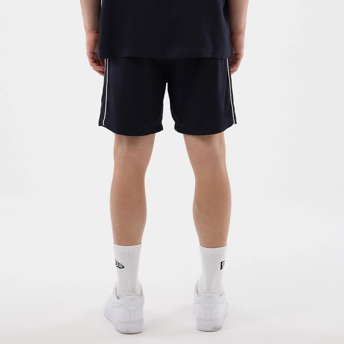 New Era Piping Navy Shorts, back view on-model showing the navy design with contrasting white side piping, ending above the knee, along with white socks featuring a New Era logo and white trainers completing the casual sporty look.