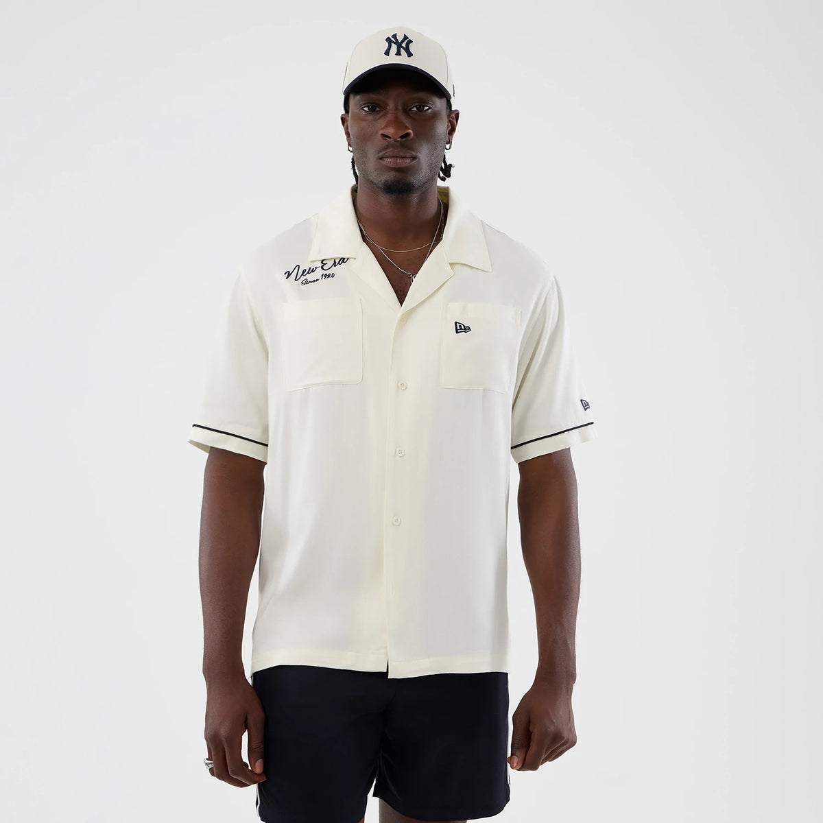 New Era Graphic Revere Open White Short Sleeve Shirt, front view on a model with a New York Yankees cap, featuring two chest pockets, the New Era script and logo on the chest, and black piping on the cuffs and collar.