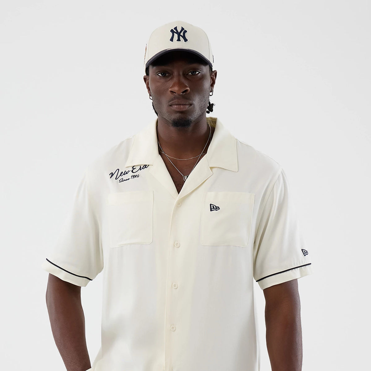 New Era Graphic Revere Open White Short Sleeve Shirt, front view on a model with an embroidered navy New Era logo over the left chest pocket, detailed with New Era Since 1920 script on the right chest, and black piping on the sleeves.