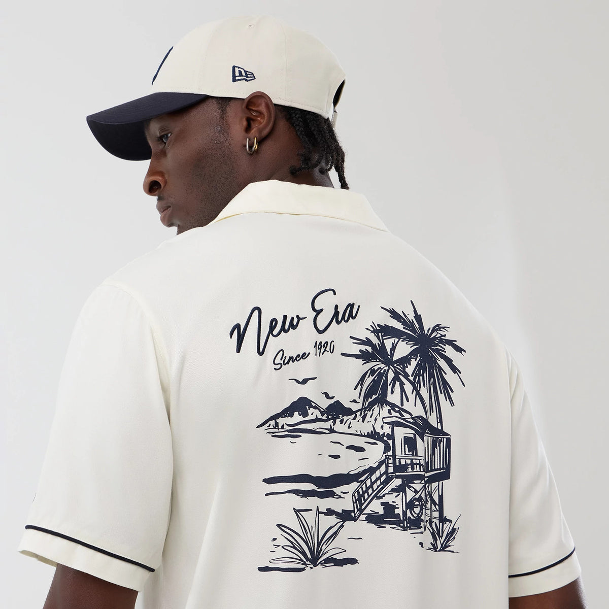 New Era Graphic Revere Open White Short Sleeve Shirt, back view on a model with a contrasting cap, featuring a navy beach scene graphic with palm trees and the New Era Since 1920 script across the upper back, enhanced with dark piping on the sleeves.