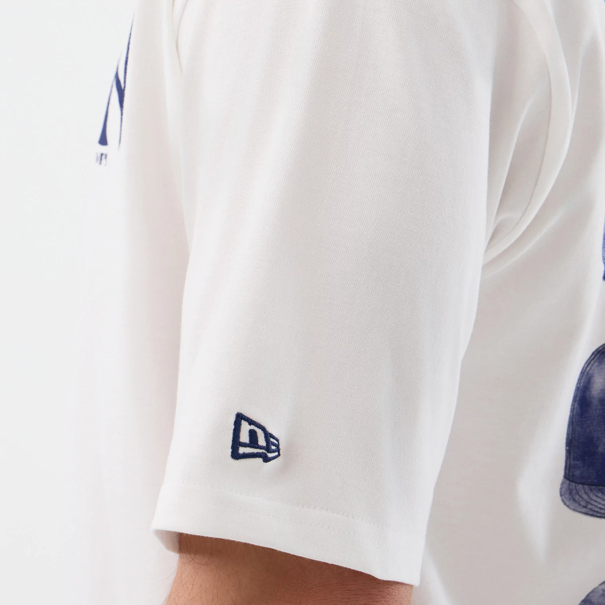 New Era New York Yankees MLB Cap Graphic White Oversized T-Shirt, side view showing the sleeve with a blue embroidered New Era logo near the hem, a view of a large cap graphic on the back, and blue lettering near the shoulder.