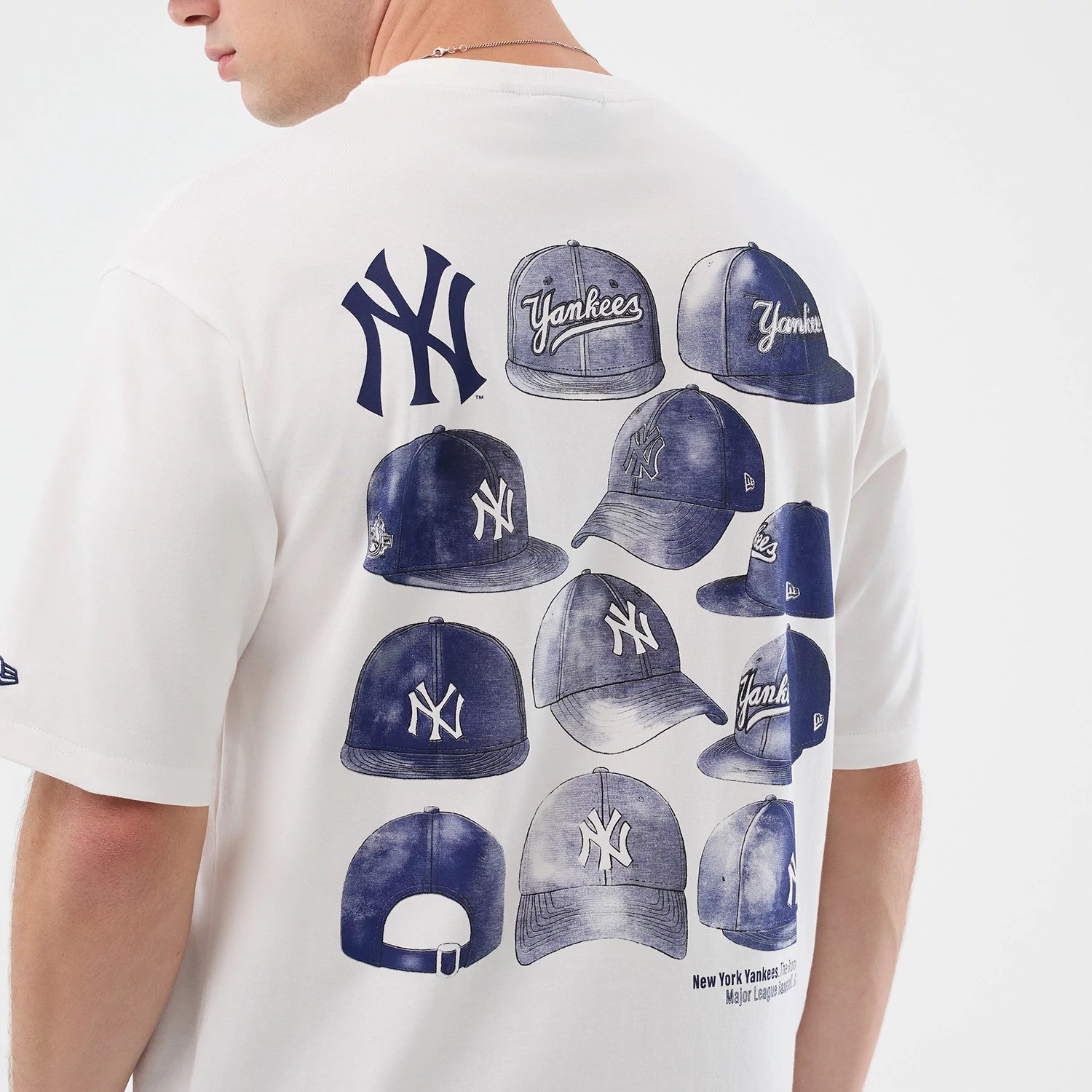 New Era New York Yankees MLB Cap Graphic White Oversized T-Shirt, back view on a model revealing a large printed design of various blue-toned Yankees caps, a prominent navy New York Yankees logo above, and a small New Era logo on the left sleeve.