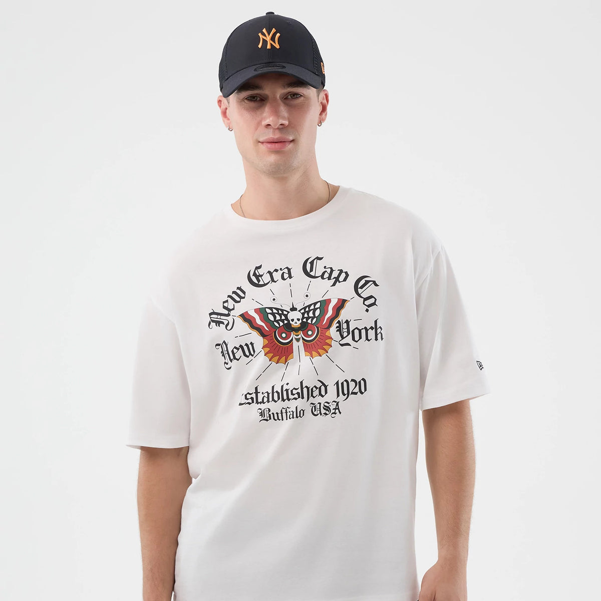 New Era Death Moth Graphic White Oversized T-Shirt, front view on a model styled with a black cap, featuring a large multicoloured moth graphic with skull and lettering, New Era Cap Co. New York printed across the chest.