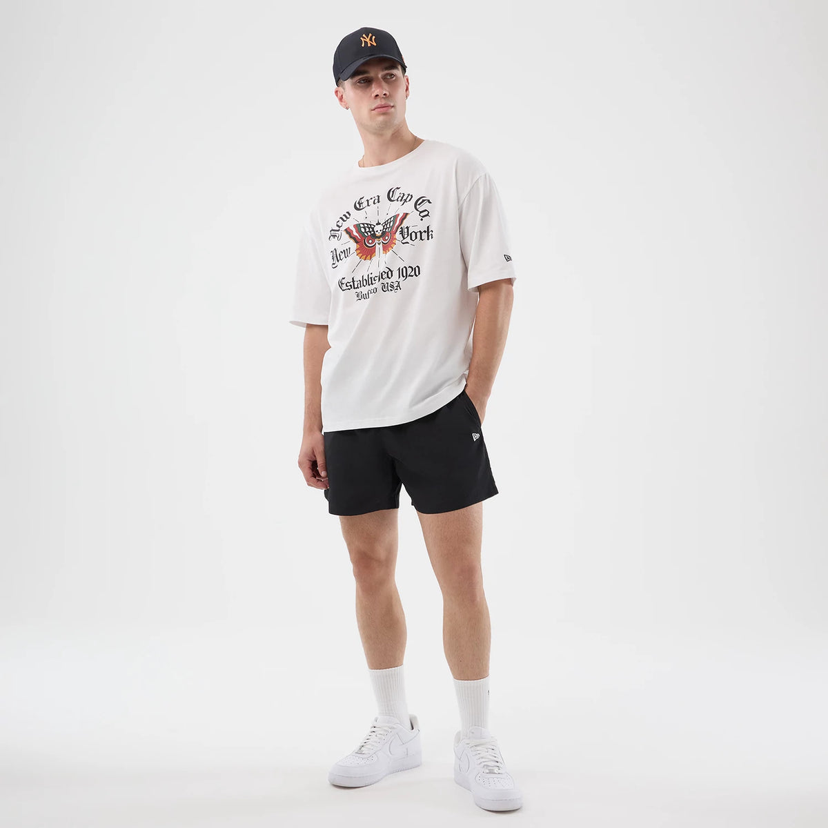 New Era Death Moth Graphic White Oversized T-Shirt, front view on a model paired with black shorts and a dark cap, featuring a central moth print with surrounding New Era Cap Co and New York text, and an embroidered New Era logo on the sleeve.