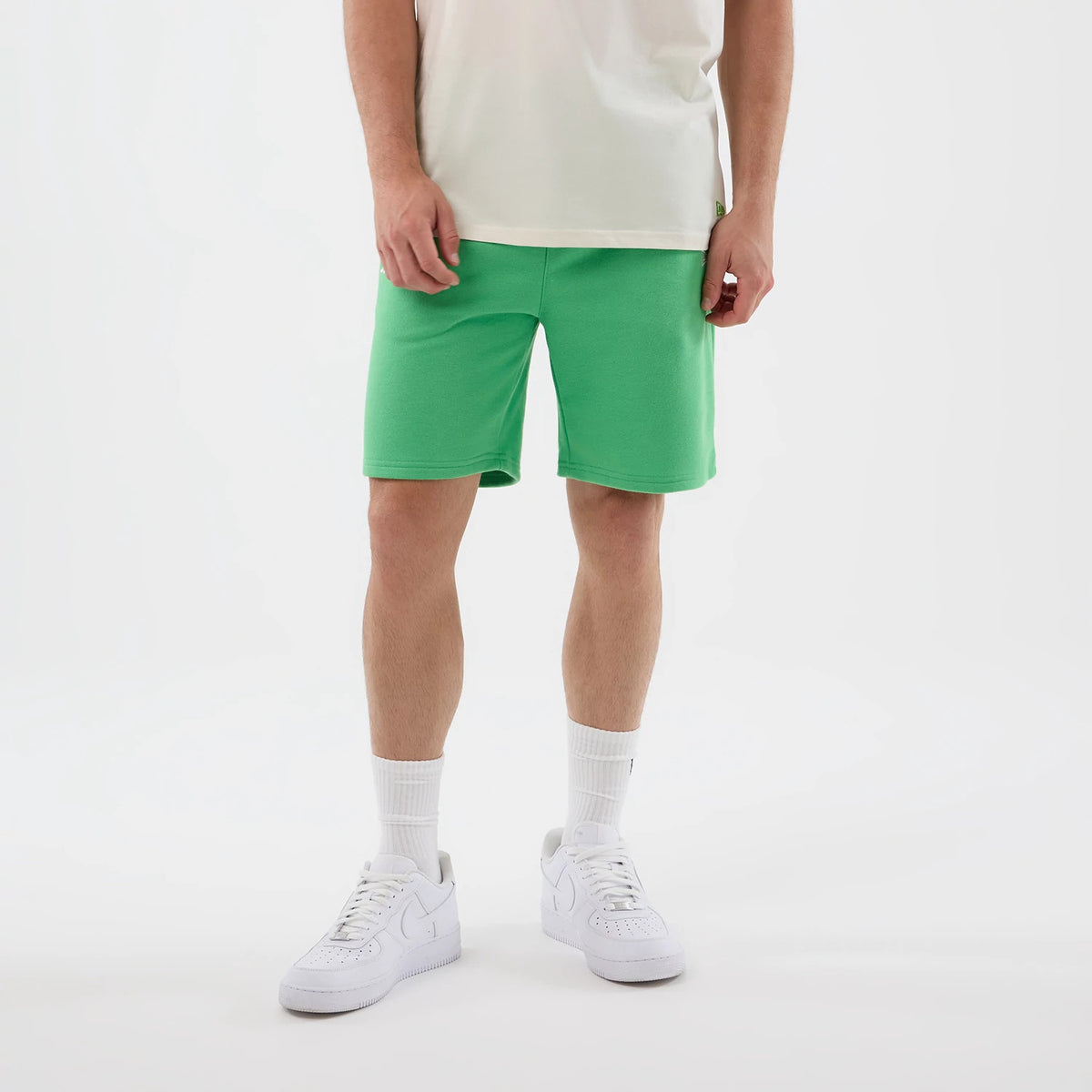 New Era LA Dodgers MLB League Essential Open Green Shorts, front view on-model showing the bright green colour and knee-length cut, paired with white trainers and socks, with a black embroidered New Era logo on the left thigh.