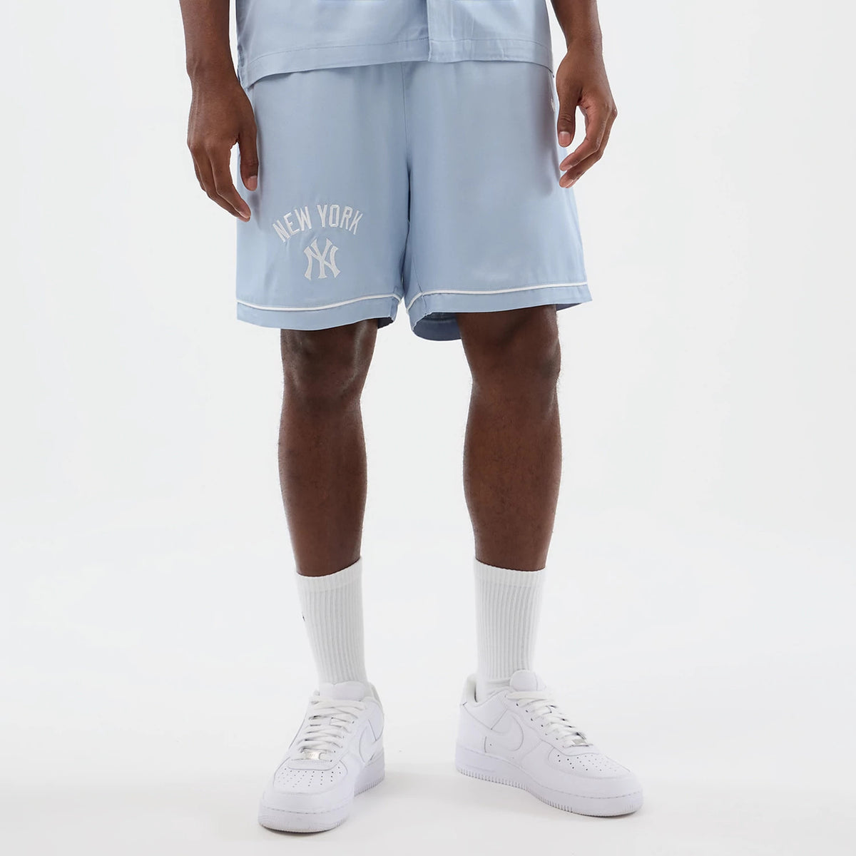 New Era New York Yankees MLB Woven Pastel Blue Shorts, front view on a model with white trainers, showcasing the embroidered New York Yankees logo and lettering on the left leg and a white trim along the hem of the light blue shorts.