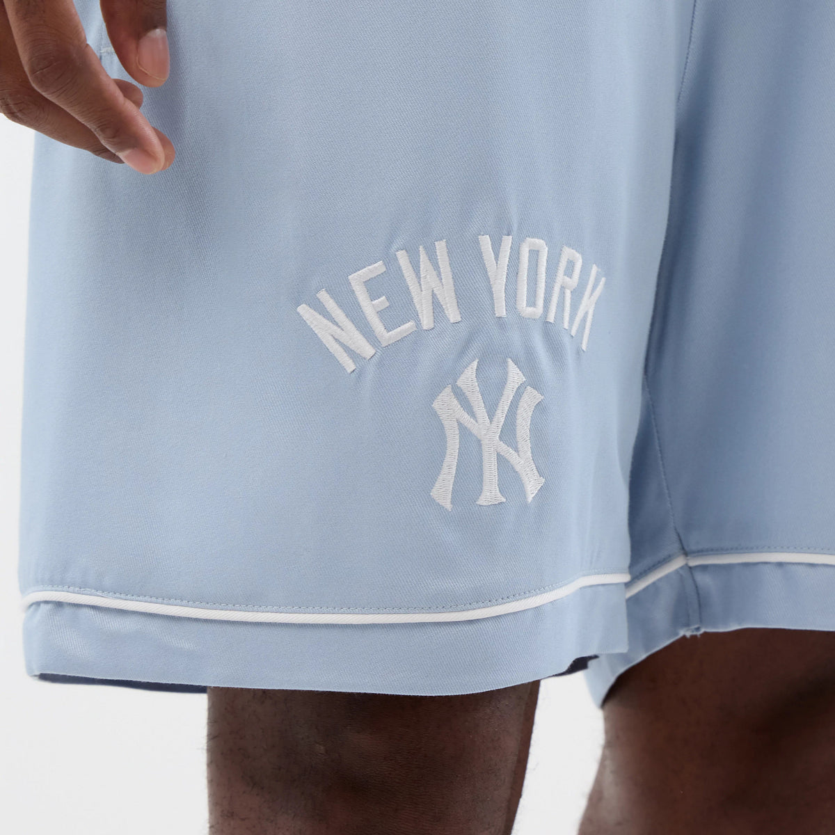 New Era New York Yankees MLB Woven Pastel Blue Shorts, front view showing an embroidered white New York Yankees logo and lettering on the left leg, with a white piping detail running along the hem of the soft blue fabric.