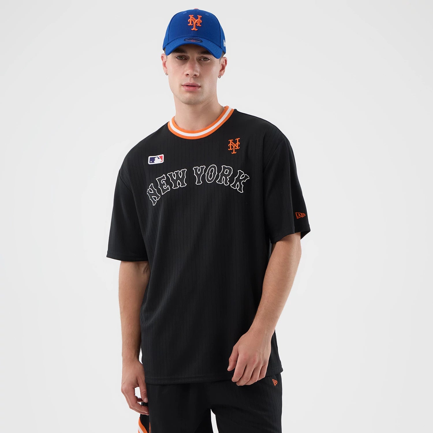 New Era New York Mets MLB Arch Black Oversized T-Shirt, front view on a model wearing a blue cap, with an embroidered orange Mets logo near the collar, New York lettering in arched white embroidery across the chest, and an orange-trimmed neckline.