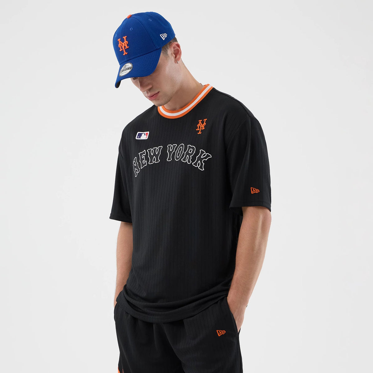 New Era New York Mets MLB Arch Black Oversized T-Shirt, front view on a model wearing a blue cap, showing orange embroidered Mets logo on the left chest, white New York lettering in arch across the chest, and orange collar trim.