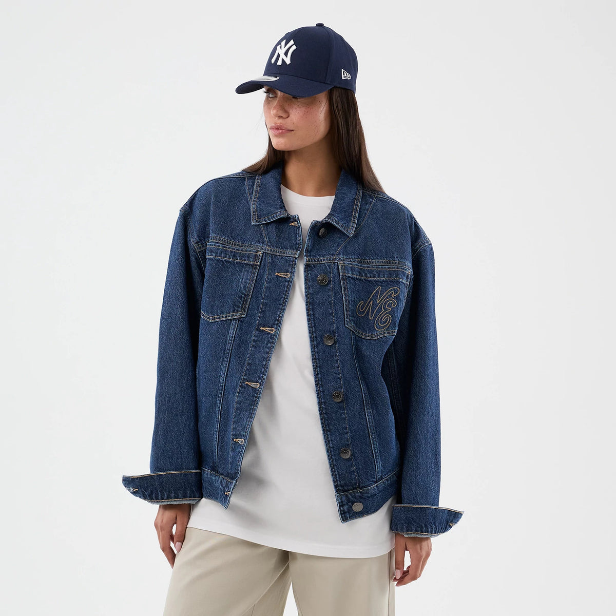 New Era Script Denim Dark Blue Jacket, front view on-model showcasing a dark blue denim jacket with large buttons, chest pockets, and an embroidered NE logo on the left chest, styled over a white top and beige trousers.