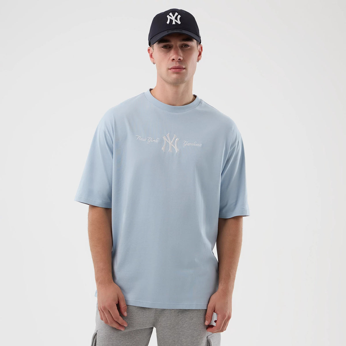 New Era New York Yankees MLB Script Pastel Blue Oversized T-Shirt, front view on a model styled with a matching navy cap, featuring an embroidered New York Yankees logo in the centre of the chest flanked by script lettering on either side.