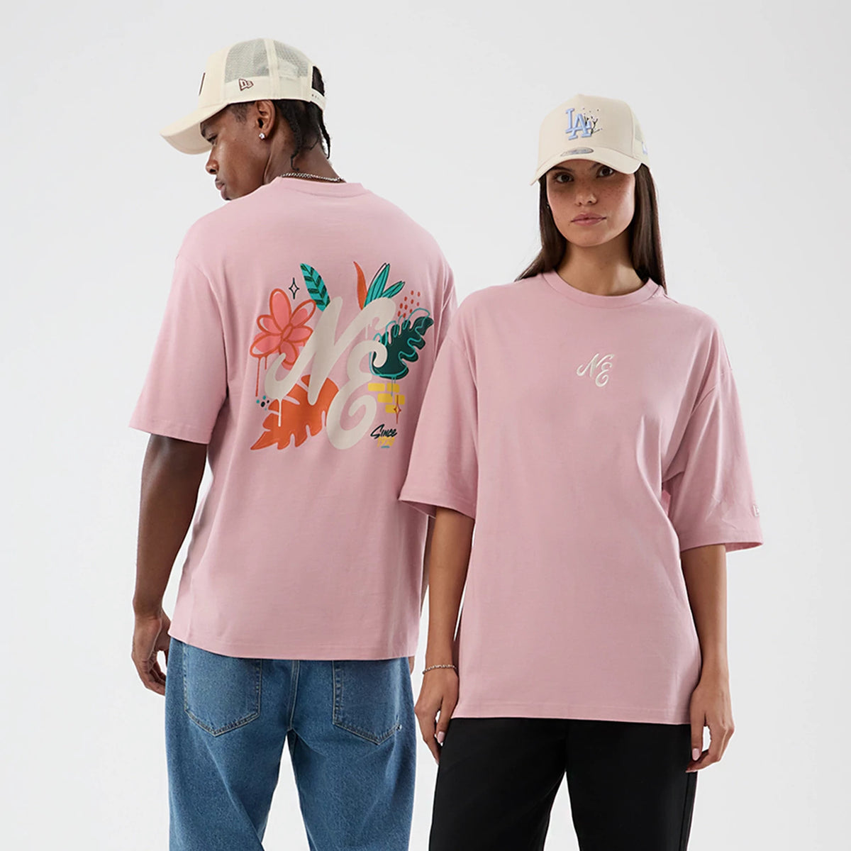 New Era Floral Graphic Pastel Pink Oversized T-Shirt, on-model view featuring one person showing the floral graphic, with NE lettering across the back, and another displaying the small NE embroidered logo on the front chest.
