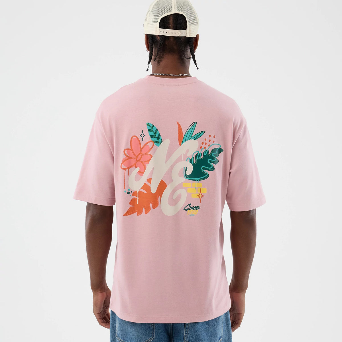 New Era Floral Graphic Pastel Pink Oversized T-Shirt, back view on a model wearing a cap, featuring a large floral design with NE lettering in cream, surrounded by colourful leaves and flowers, and a small Since detail near the bottom.