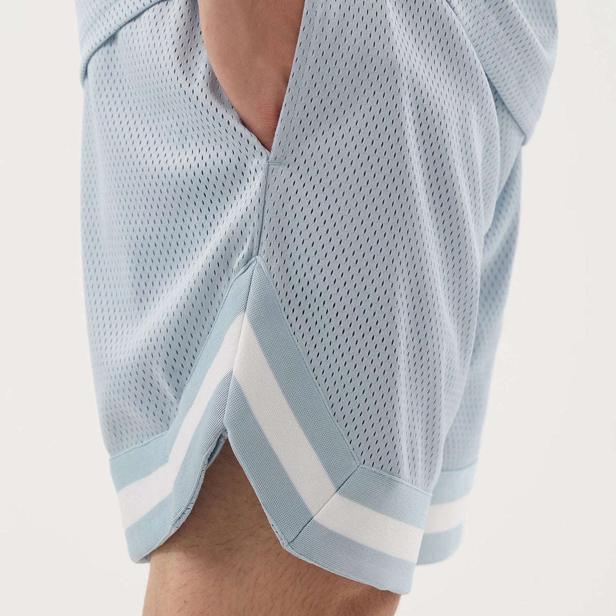 New Era Mesh Pastel Blue Oversized Shorts, on-model view showing the dotted mesh texture, a side pocket, and the white stripes along the lower hem, with a distinct side slit for added style on the light blue fabric.