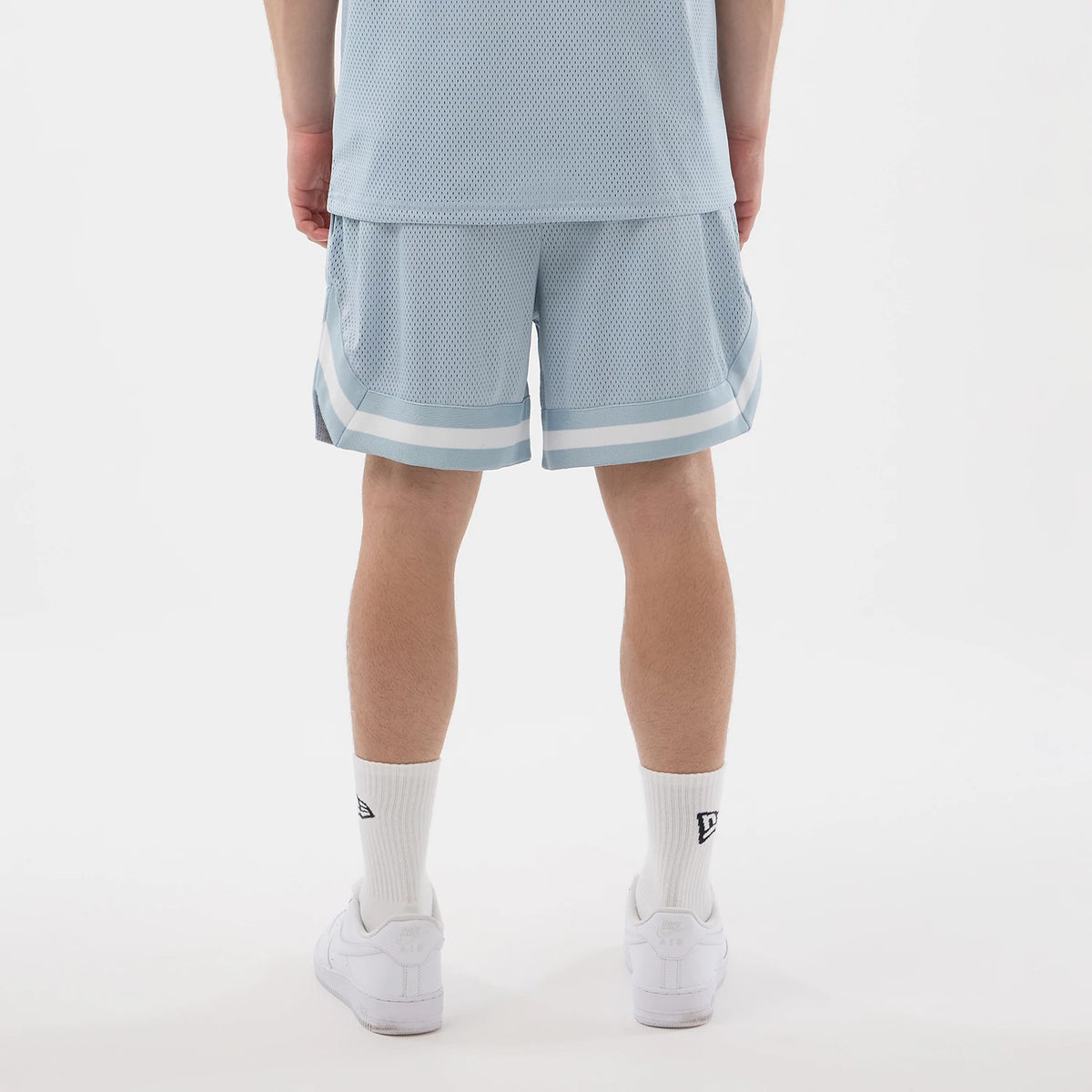 New Era Mesh Pastel Blue Oversized Shorts, back view showing the loose fit with light blue and white horizontal stripes at the hem, mesh texture throughout, styled with a matching top, white socks, and trainers.