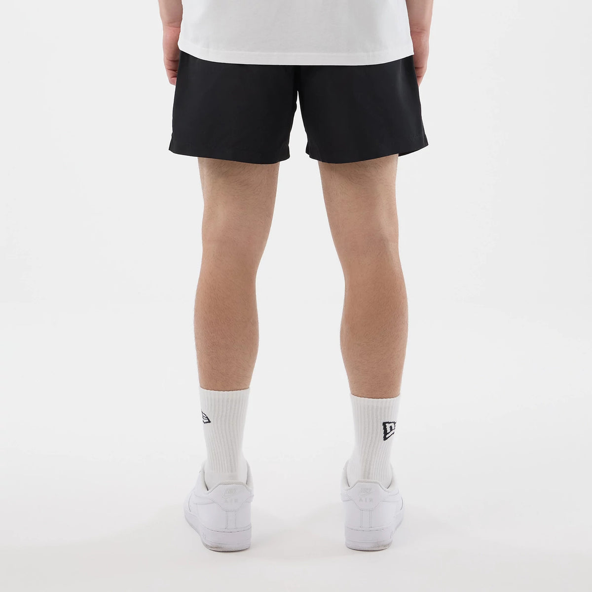 New Era Black Swimming Shorts, back view on a model showing the simple black design reaching mid-thigh, complemented by plain white socks featuring embroidered logos, and white trainers that add a casual touch to the overall look.