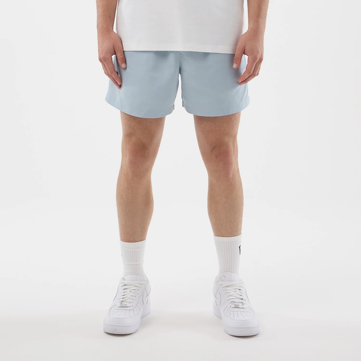 New Era Pastel Blue Swimming Shorts, front view on-model showcasing a simple and clean design in a soft light blue colour reaching mid-thigh, paired with a white T-shirt and white sneakers for a casual summer look.