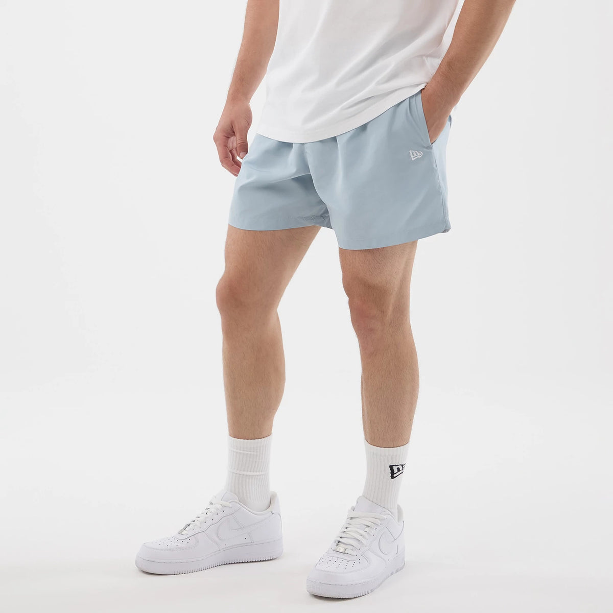 New Era Pastel Blue Swimming Shorts, front view on-model showing a light blue colour with relaxed fit, side pockets, and an embroidered New Era logo on the left leg, styled with white trainers and crew socks.