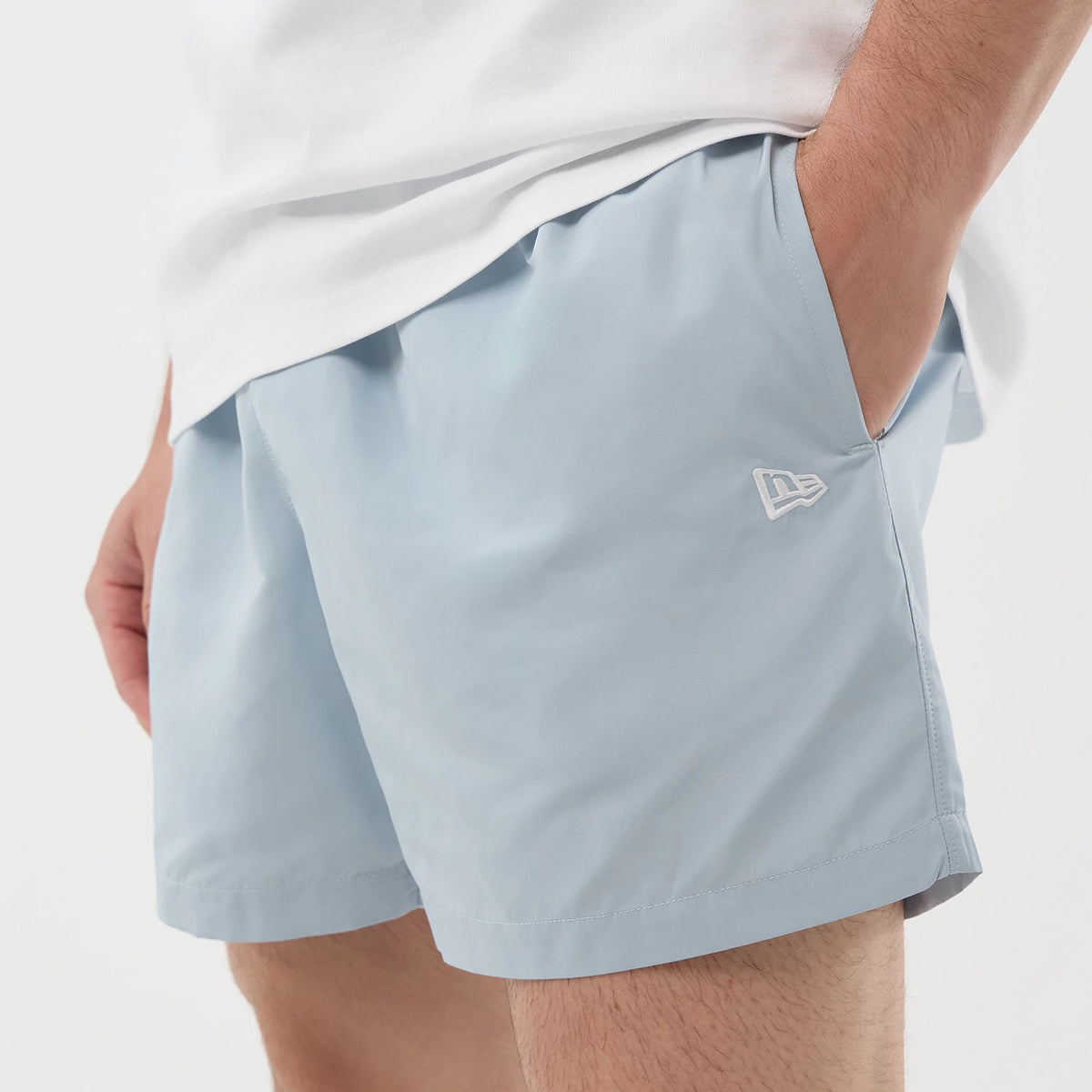 New Era Pastel Blue Swimming Shorts, front view on a model showcasing the light blue colour with a hemline above the knee, an embroidered white New Era logo on the left leg, and a relaxed fit paired with a plain white shirt.