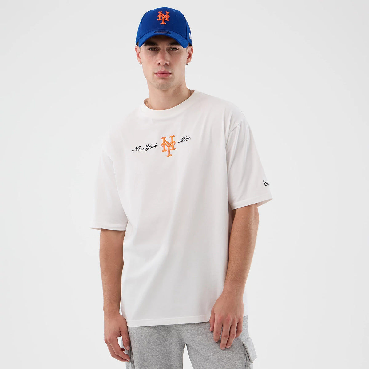 New Era New York Mets MLB Script White Oversized T-Shirt, front view on-model showing the orange embroidered Mets logo centred on the chest with black New York Mets script on either side, complemented by a matching blue cap and grey shorts.