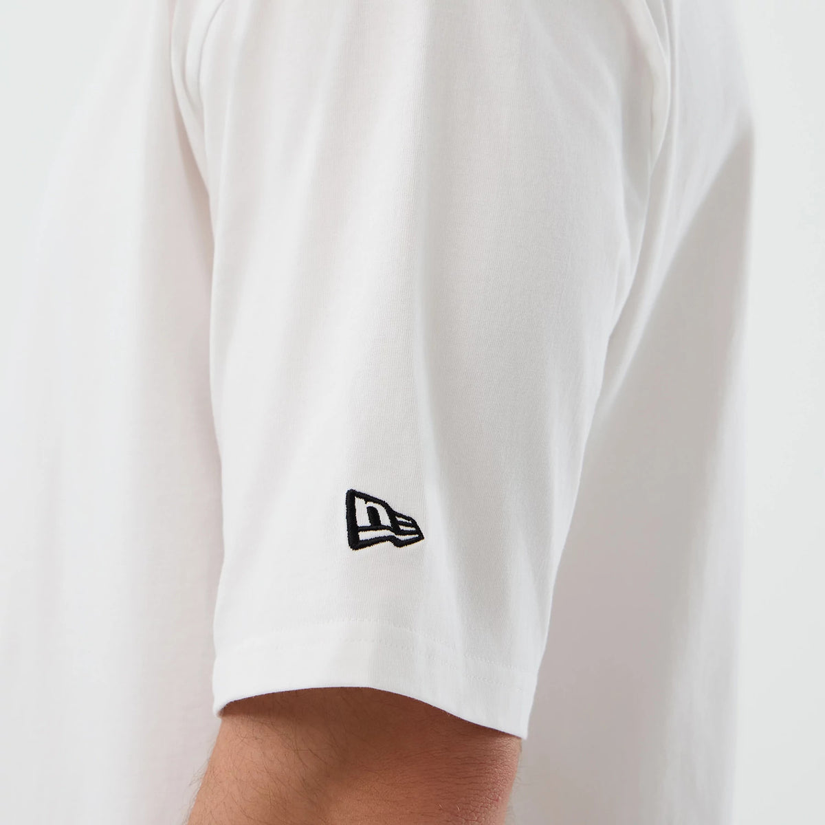 New Era New York Mets MLB Script White Oversized T-Shirt, side view focusing on the short sleeve with a black embroidered New Era flag logo near the hem, showing the clean white fabric and the drape over the arm.