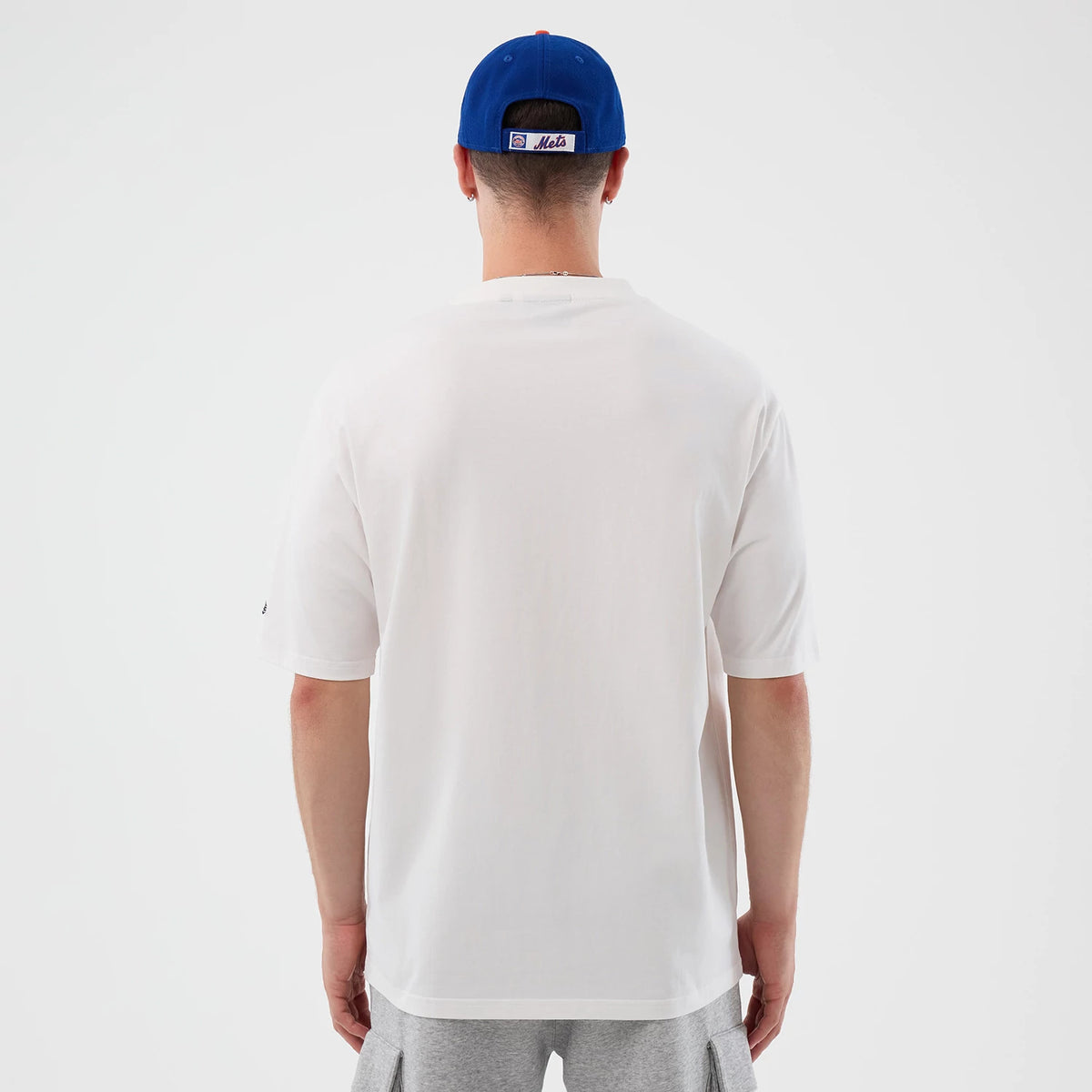 New Era New York Mets MLB Script White Oversized T-Shirt, back view on a model styled with a blue cap, displaying a clean white back and sleeves, a small embroidered New Era logo on the left sleeve, and a loose fit extending to grey shorts.