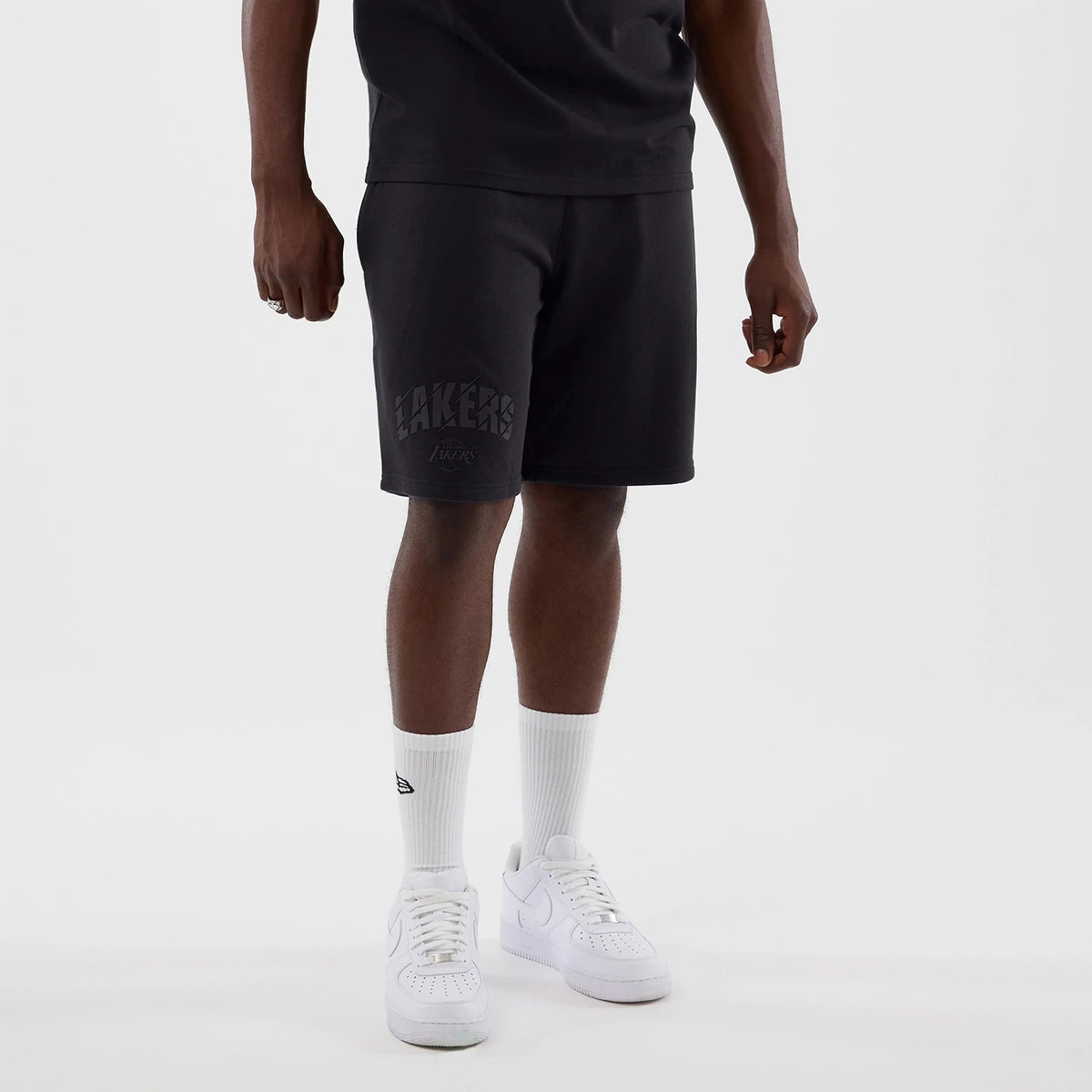 New Era LA Lakers NBA Core Plus Navy Shorts, front view on a model with white trainers and socks, displaying a black embroidered Lakers logo on the left leg, an elastic waistband at the top and a plain navy fabric throughout.