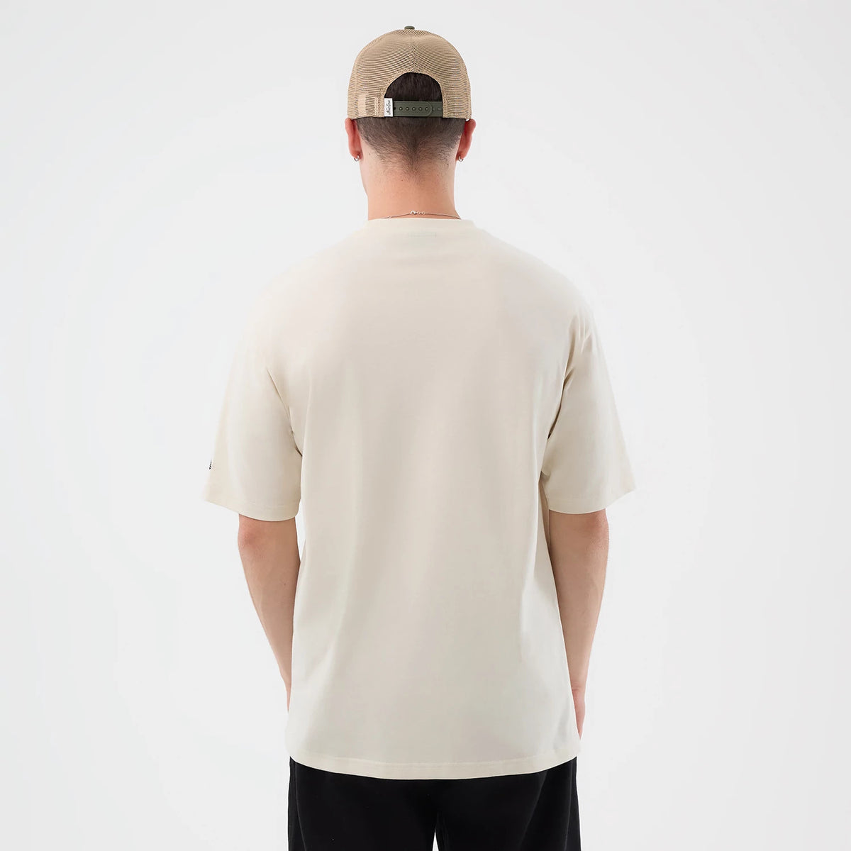 New Era Dessert Graphic Light Beige Oversized T-Shirt, back view on a model wearing a beige cap, showing the plain light beige fabric extending over the shoulders and short sleeves, with a loose fit and straight hemline.