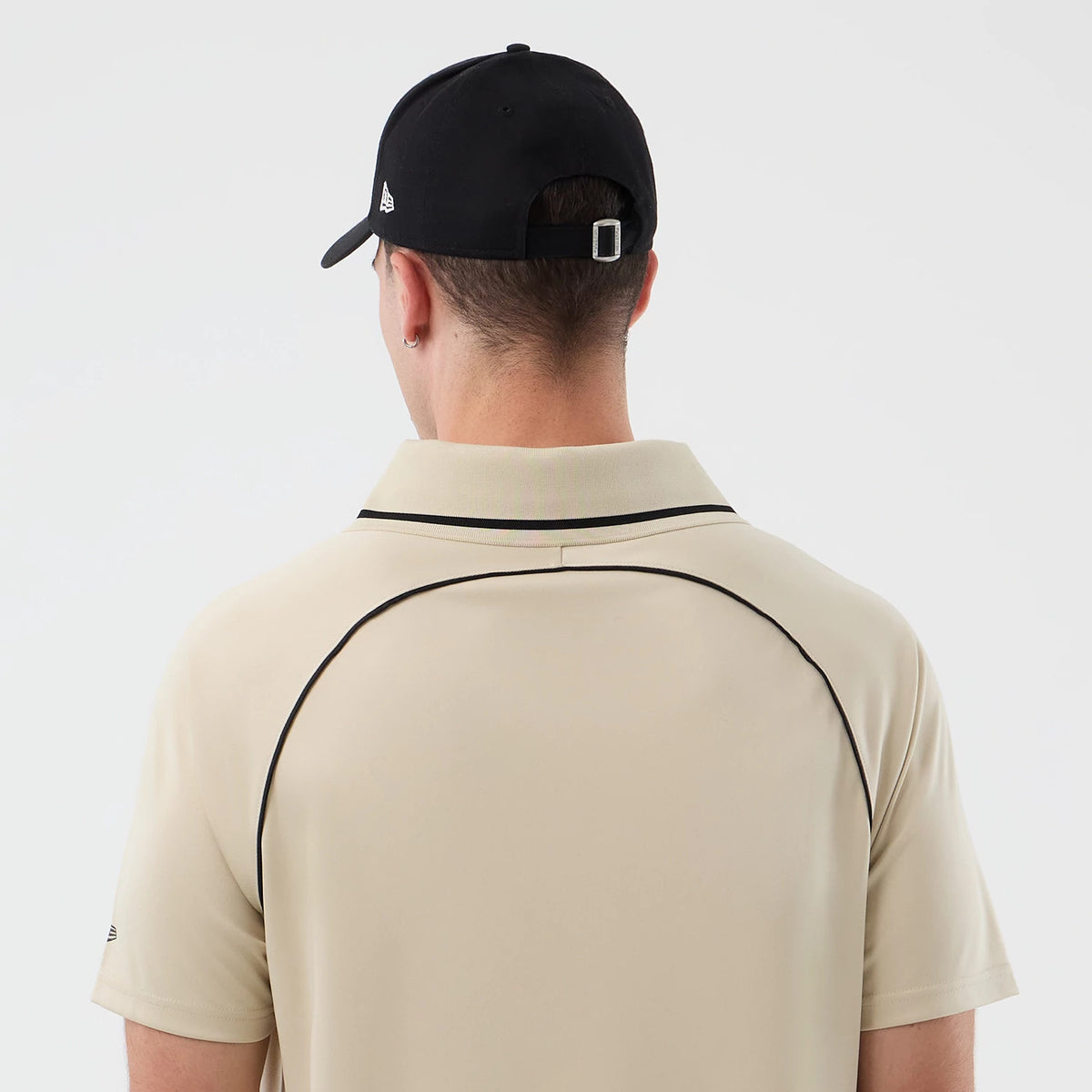New Era Football Light Beige T-Shirt, back view on a model wearing a black cap, showing contrasting black piping along the shoulders and sleeve edges, an embroidered New Era logo on the left sleeve, and a folded collar.