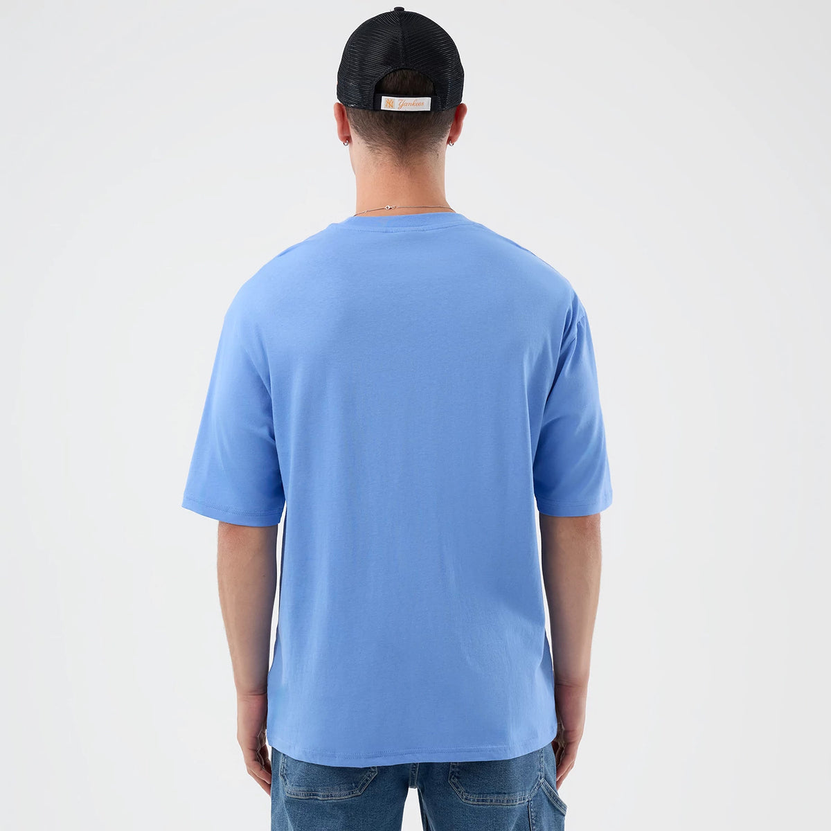New Era Basketball Character Graphic Pastel Blue T-Shirt, back view on a model showcasing the plain light blue back with short sleeves, paired simply with jeans and a black cap for a casual look.