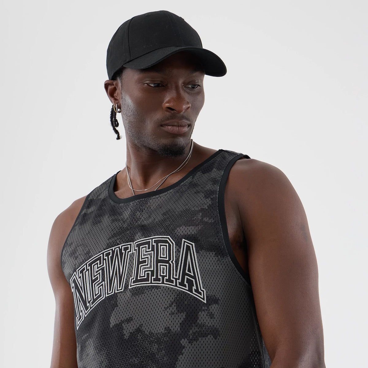 New Era All Over Print Black Camo Mesh Tank Top, on-model front view with a mix of black and grey camo print, featuring large outlined New Era lettering across the chest, and paired with a plain black cap.