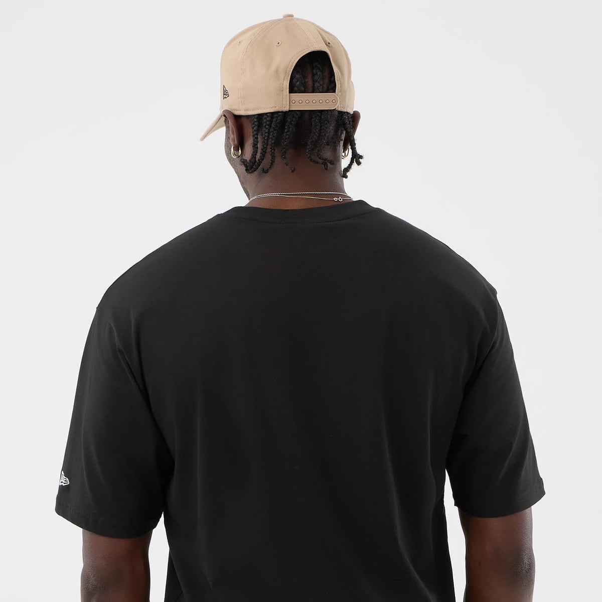 New Era Serpent Graphic Black Oversized T-Shirt, back view on a model wearing a beige cap, featuring a plain black back with short sleeves and a small embroidered New Era logo on the left sleeve near the hem.