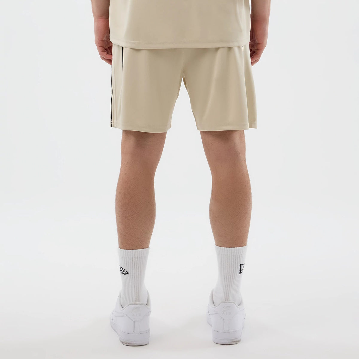 New Era Football Light Beige Shorts, back view on a model wearing matching light beige shorts paired with white crew socks featuring the embroidered New Era logo and white trainers, showing a straight hem just above the knee.