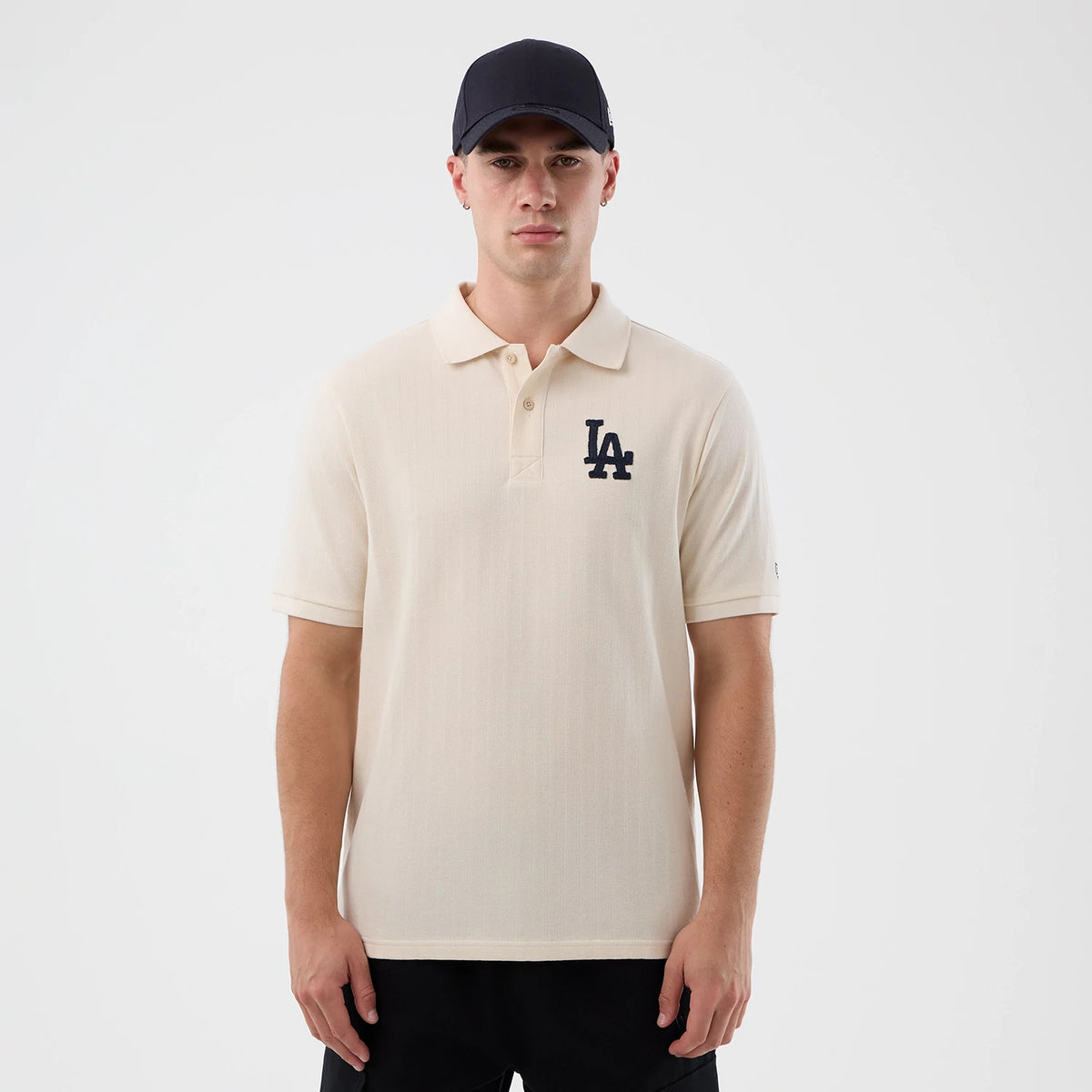 New Era LA Dodgers MLB Rib Open White Polo T-Shirt, front view on-model showcasing a light-coloured polo with a dark embroidered LA Dodgers logo on the left chest, a classic collar with a buttoned placket, and short sleeves paired with dark trousers.
