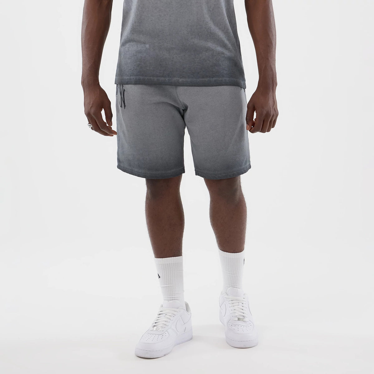 New Era New York Yankees MLB Washed Logo Black Shorts, front view on-model showing the shorts with a gradient from grey to black, a small printed Yankees logo on the left leg near the waistband, styled with white trainers and socks.