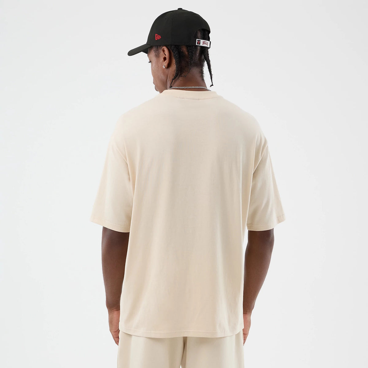 New Era Chicago Bulls NBA Core Plus Light Beige Oversized T-Shirt, back view on a model wearing a black cap, highlighting the plain light beige fabric extending over the shoulders and upper back, showcasing the relaxed fit with short sleeves.