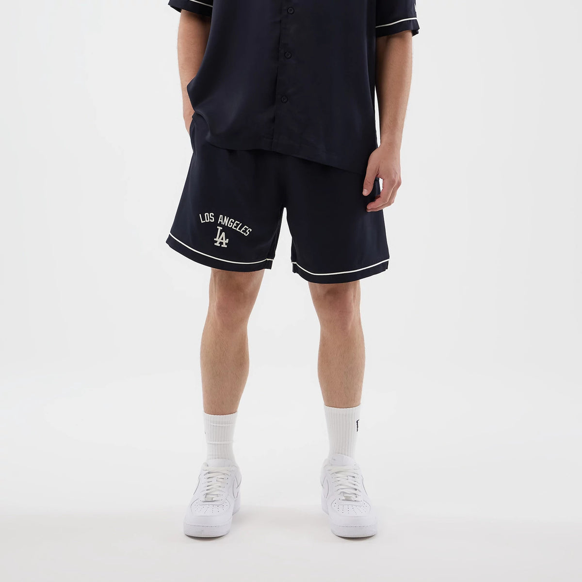New Era LA Dodgers MLB Woven Navy Shorts, front view on a model showing the embroidered Los Angeles lettering and LA logo on the left leg, white piping along the edges, and paired with a dark button-up shirt and white trainers.