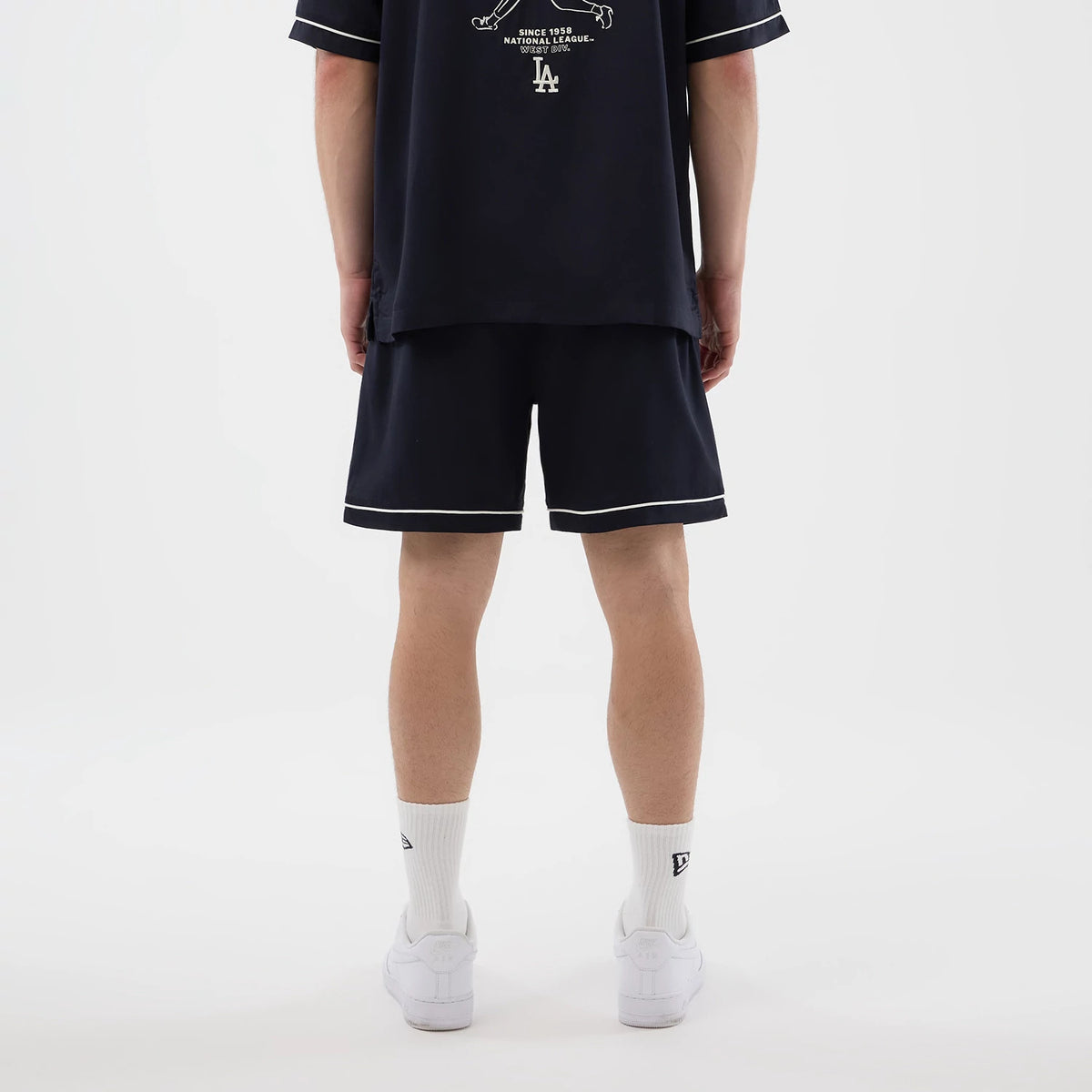New Era LA Dodgers MLB Woven Navy Shorts, back view on a model with white trim along the hem, paired with a matching navy t-shirt featuring the Dodgers logo on the lower back, white socks, and white trainers.