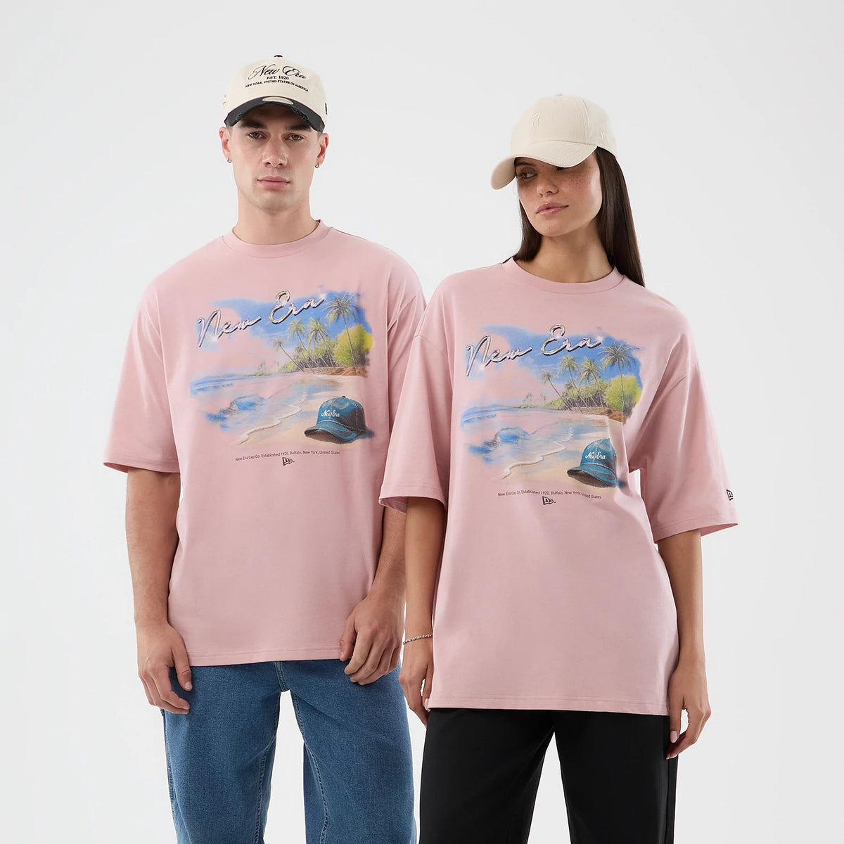 New Era Beach Graphic Pastel Pink Oversized T-Shirt, front view on a model with a neutral cap, showcasing large beach scene print with palm trees and a hat graphic, New Era script across the chest, and a small embroidered logo on the sleeve.