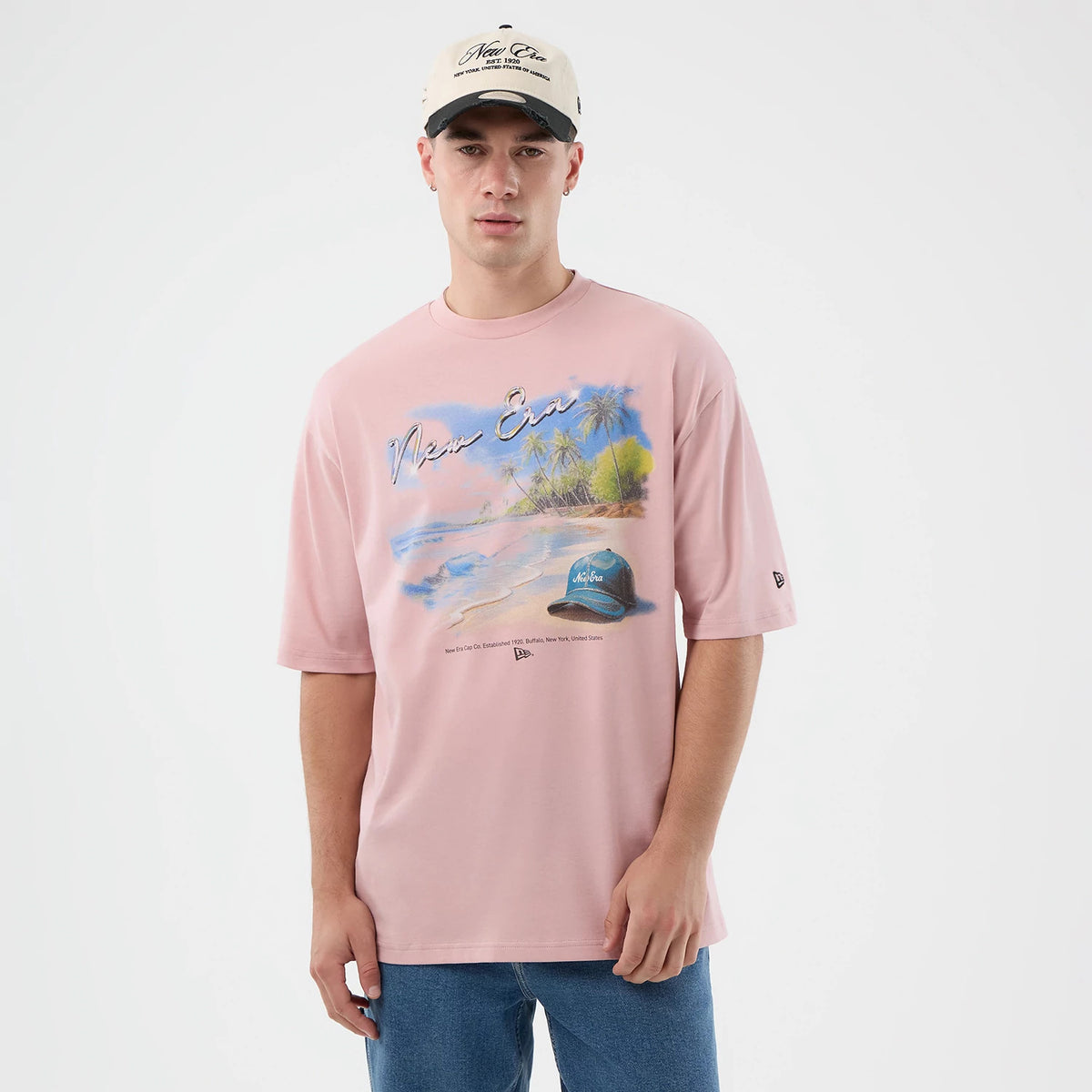 New Era Beach Graphic Pastel Pink Oversized T-Shirt, front view on a model showing a beach scene with palm trees and a blue cap printed across the chest, New Era embroidered logo on the left sleeve, styled with a beige cap and blue jeans.