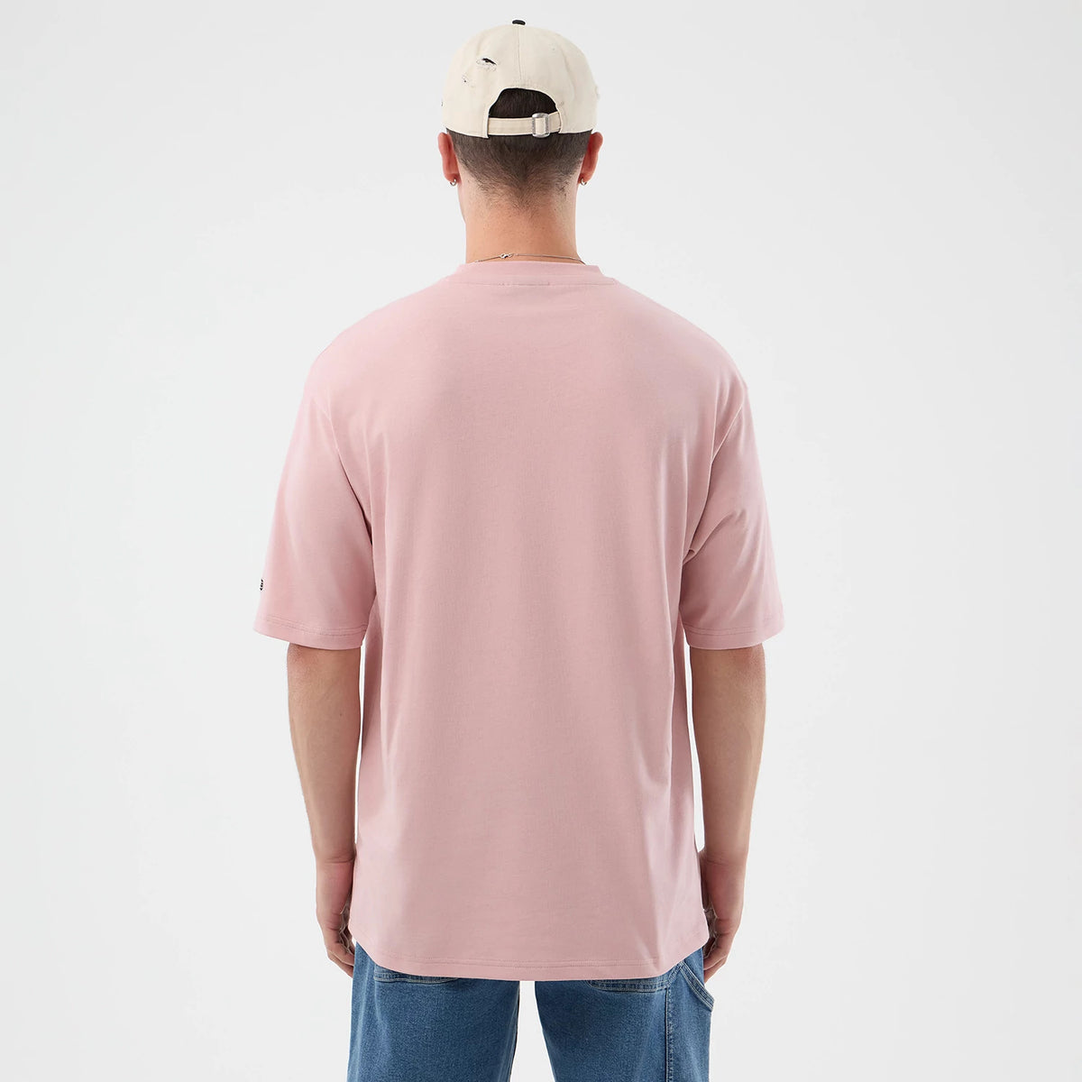 New Era Beach Graphic Pastel Pink Oversized T-Shirt, back view on a model with loose blue jeans and a beige cap, showcasing the plain pastel pink fabric and a small embroidered New Era logo on the left sleeve near the hem.