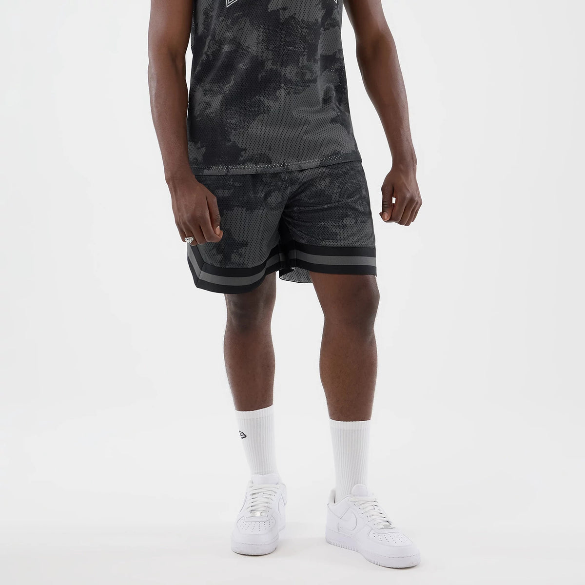 New Era All Over Print Mesh Black Camo Shorts, front view on-model showing the camouflage pattern throughout the mesh fabric, black horizontal stripes along the hem, and styling with a matching camo tank top and white trainers with long white socks.